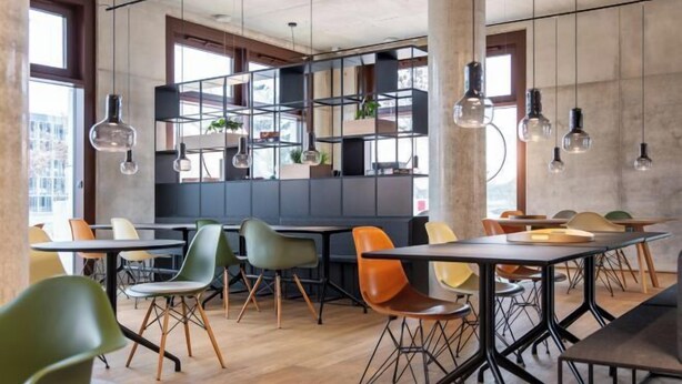 Un espace de salle à manger moderne, doté de plusieurs tables et chaises, d’une bibliothèque centrale agrémentée de plantes et de luminaires suspendus.