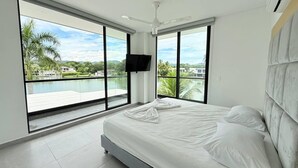 A modern bedroom with a large bed, a TV mounted on the wall, and a balcony with a pool view.