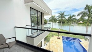 A modern balcony with a glass railing overlooking a swimming pool and a body of water with palm trees.