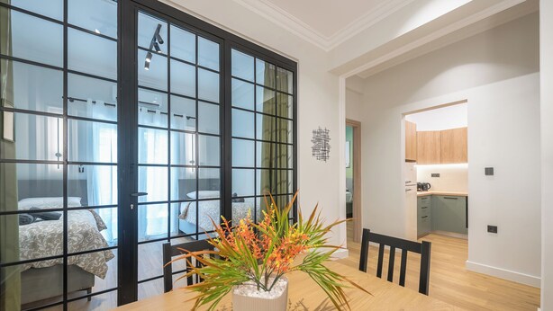 Una habitación de hotel moderna con un gran ventanal corredero que da a un balcón, un comedor con mesa y sillas de madera, y una kitchenette pequeña.