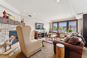 A living room with a stone fireplace, a comfortable armchair, a leather sofa, a flat-screen TV, and a large window with a view of the outside. - Steps to Park City Mtn Lift, Pool + Hot Tub, Dog Friendly, Walk to Cafe (Park City)