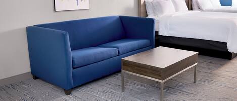 A hotel room with a blue sofa, a wooden coffee table, and a bed with white bedding.