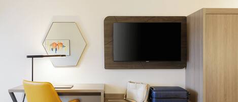 A hotel room with a desk, a yellow chair, a television, a wardrobe, and a bed.