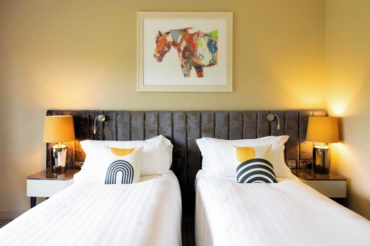 A hotel room with two beds, a large abstract painting on the wall, and lamps on either side of the beds.