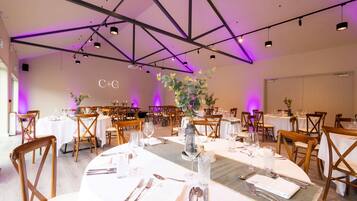 Un comedor espacioso con varias mesas dispuestas para un evento formal, que cuenta con un techo alto con vigas a la vista e iluminación colgante.