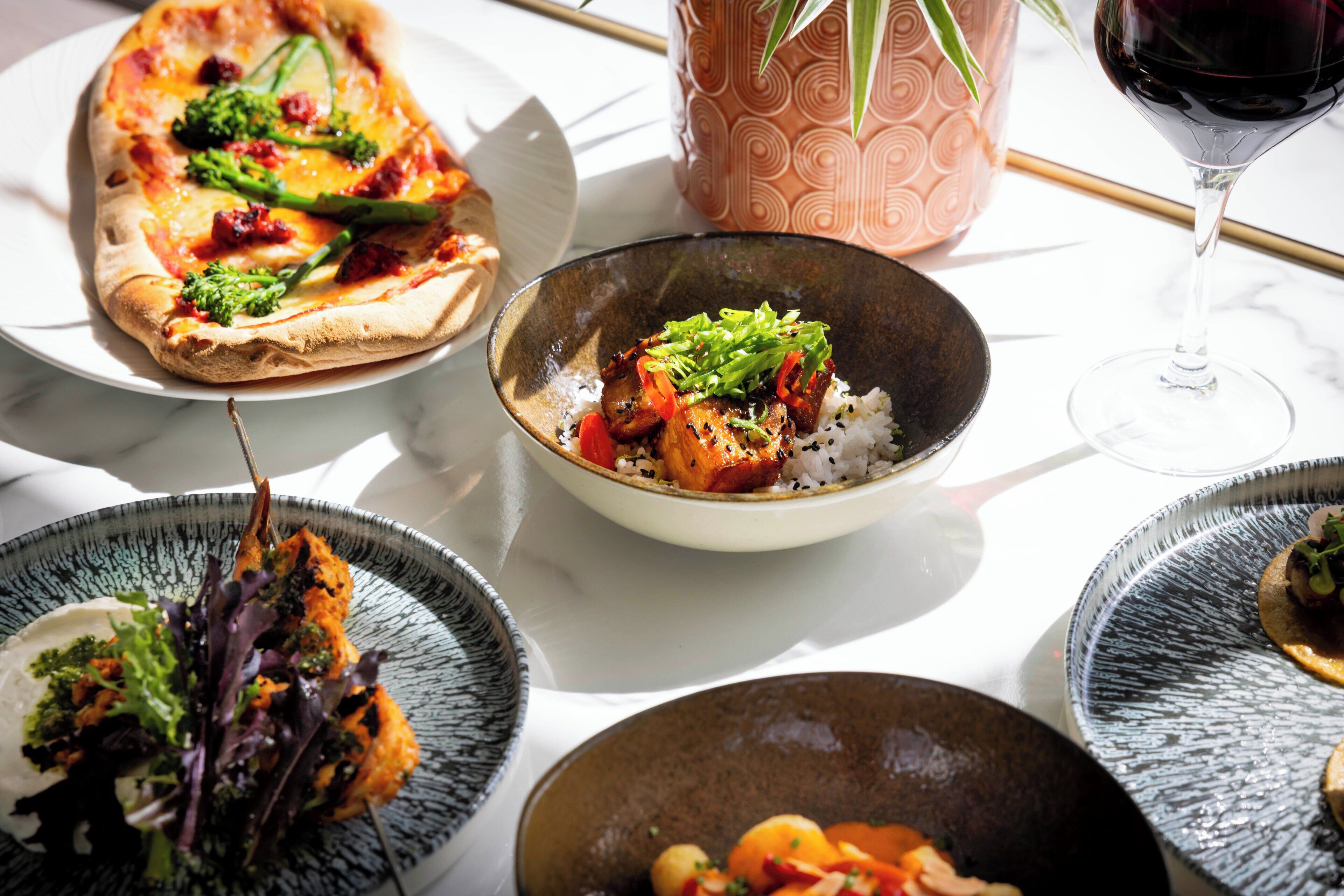 A table with a variety of dishes including a pizza, a bowl of rice with vegetables, and a glass of red wine.