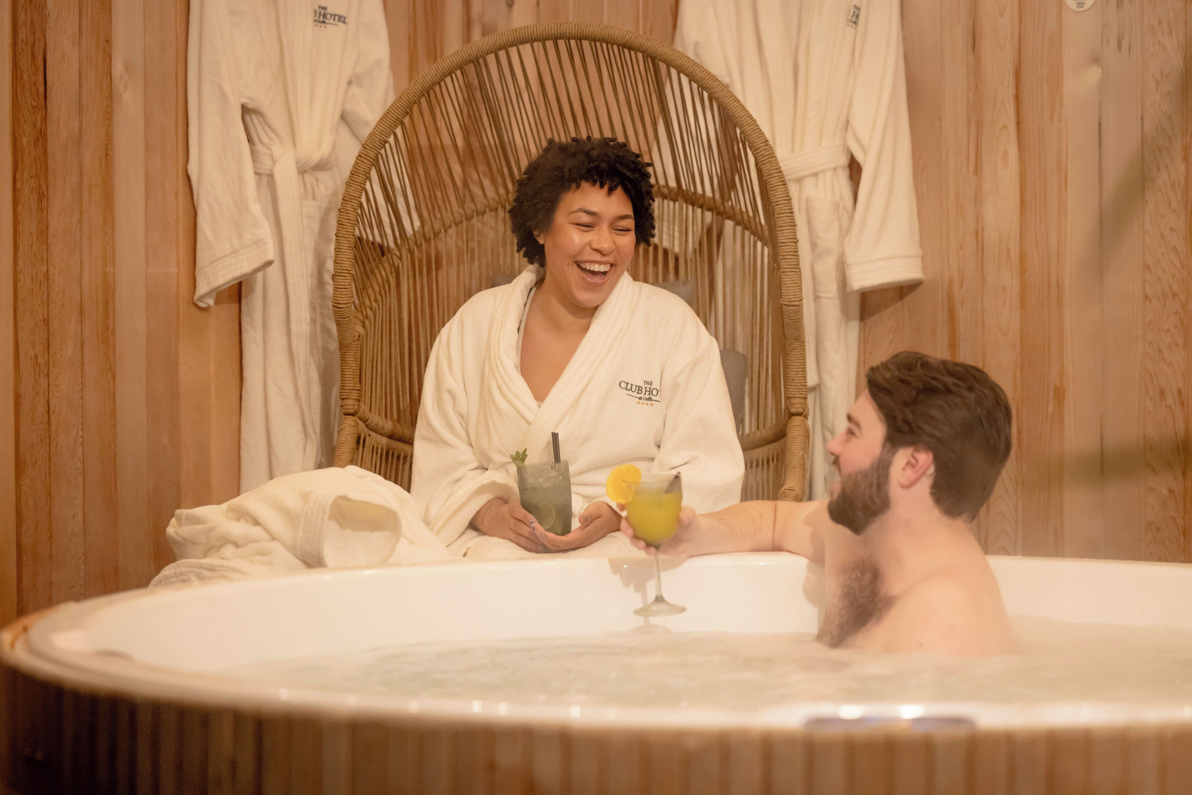 Um casal em uma banheira de hidromassagem, com interior de madeira e roupões brancos pendurados na parede.