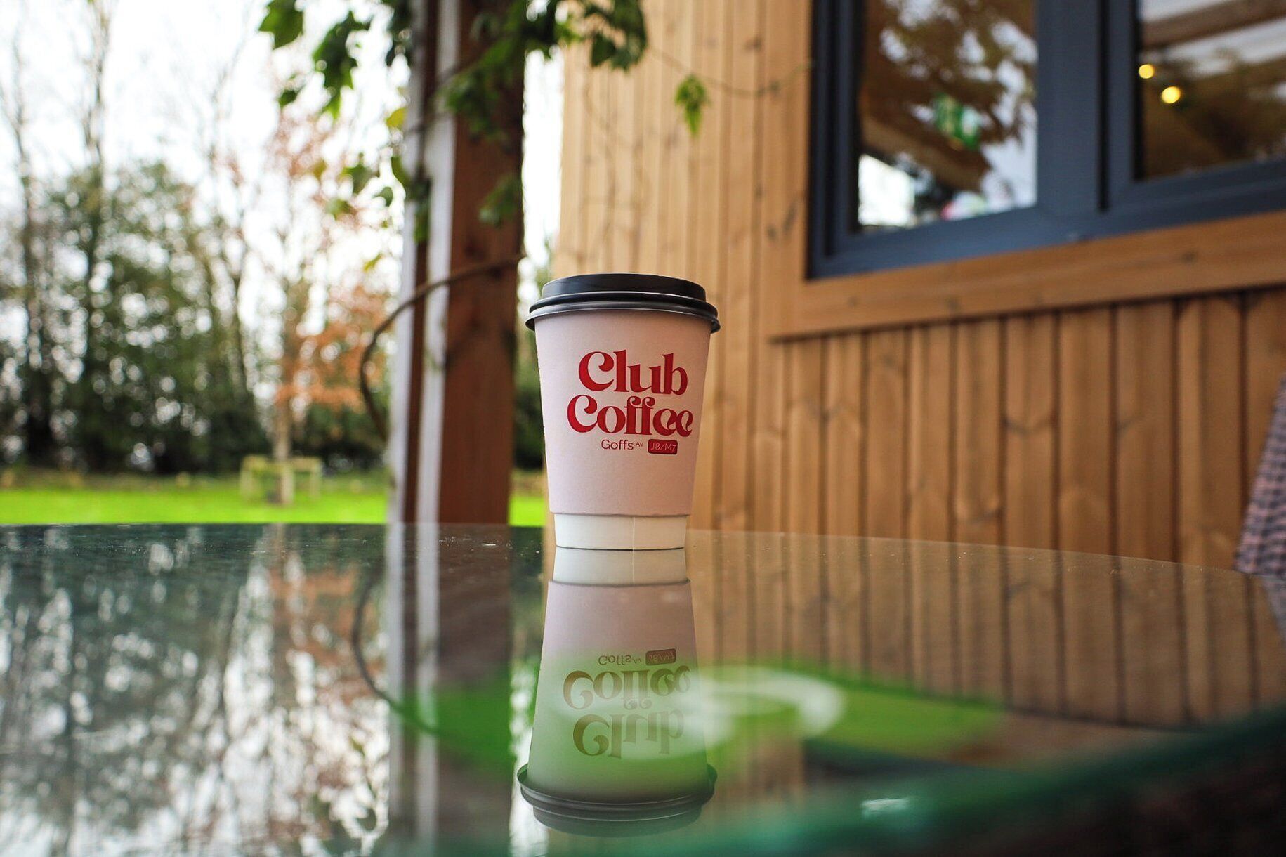 Une tasse de café Club Coffee posée sur une table en verre, avec une clôture en bois et de la verdure en arrière-plan.