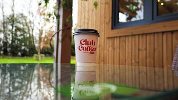 Une tasse de café Club Coffee posée sur une table en verre, avec une clôture en bois et de la verdure en arrière-plan.