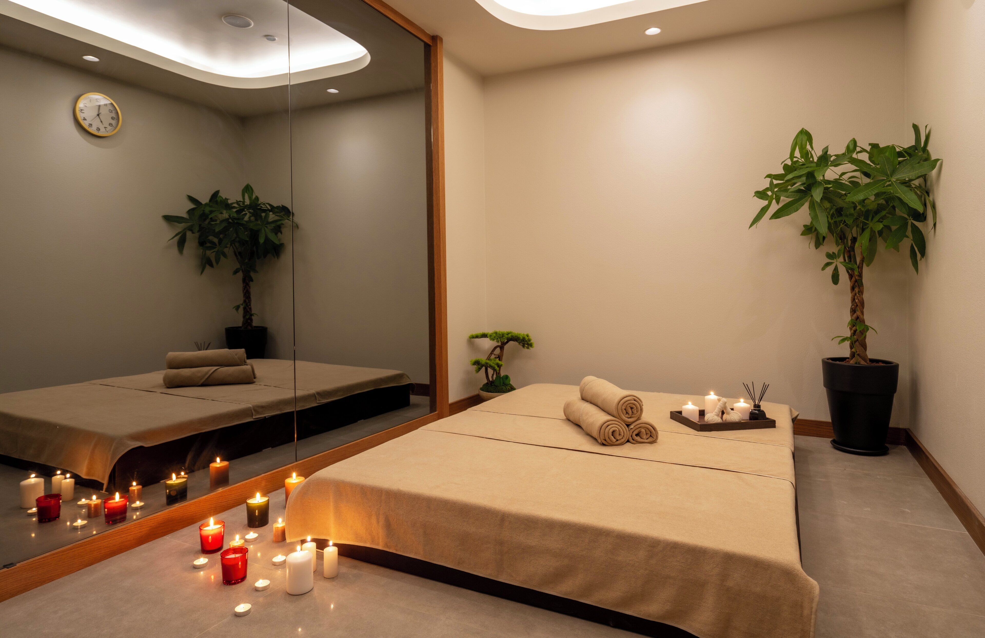 A spa room with a bed, candles, a clock, and two potted plants.