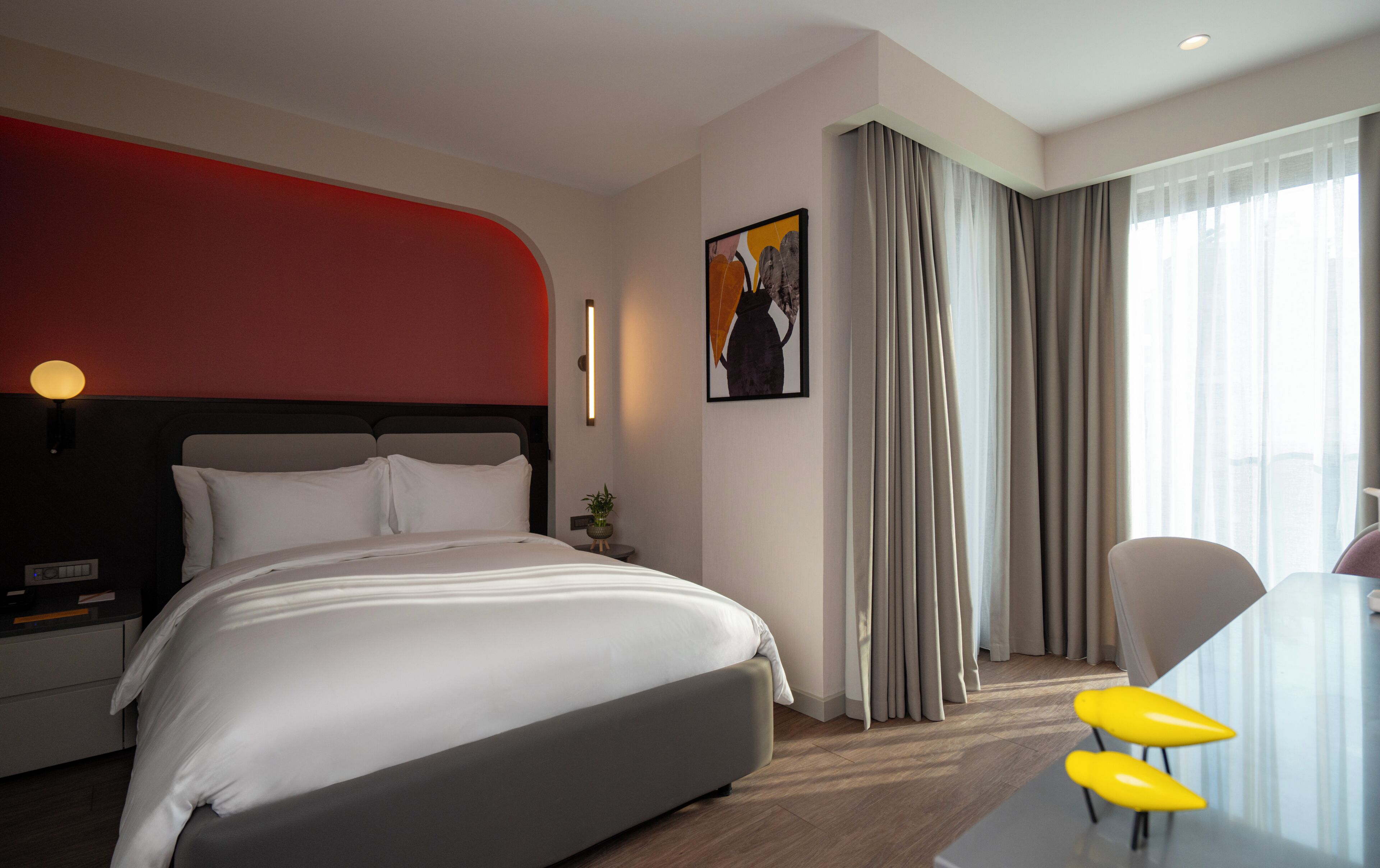 Habitación de hotel con cama, mesita de noche, lámpara, un cuadro, ventana con cortinas y una silla.