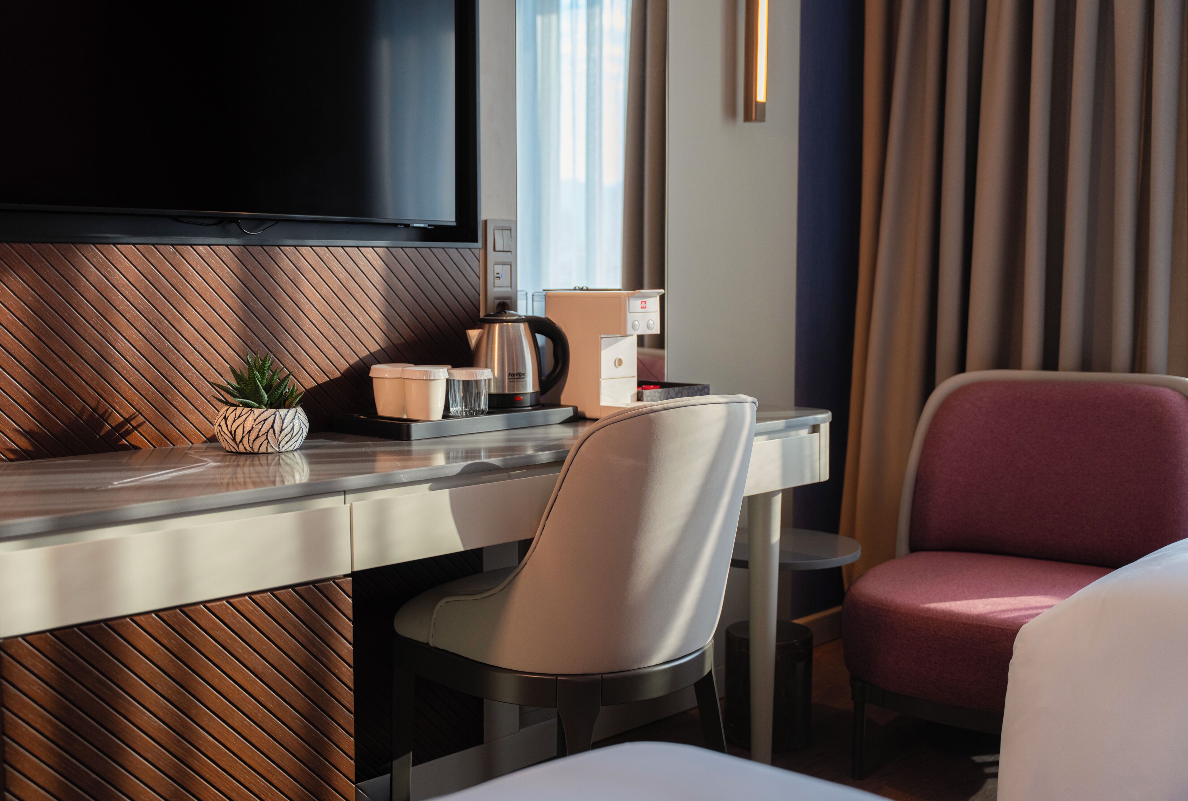 A modern hotel room with a flat-screen TV mounted on the wall, a wooden accent wall, a small potted plant, a coffee maker, and a seating area with a chair and a couch.