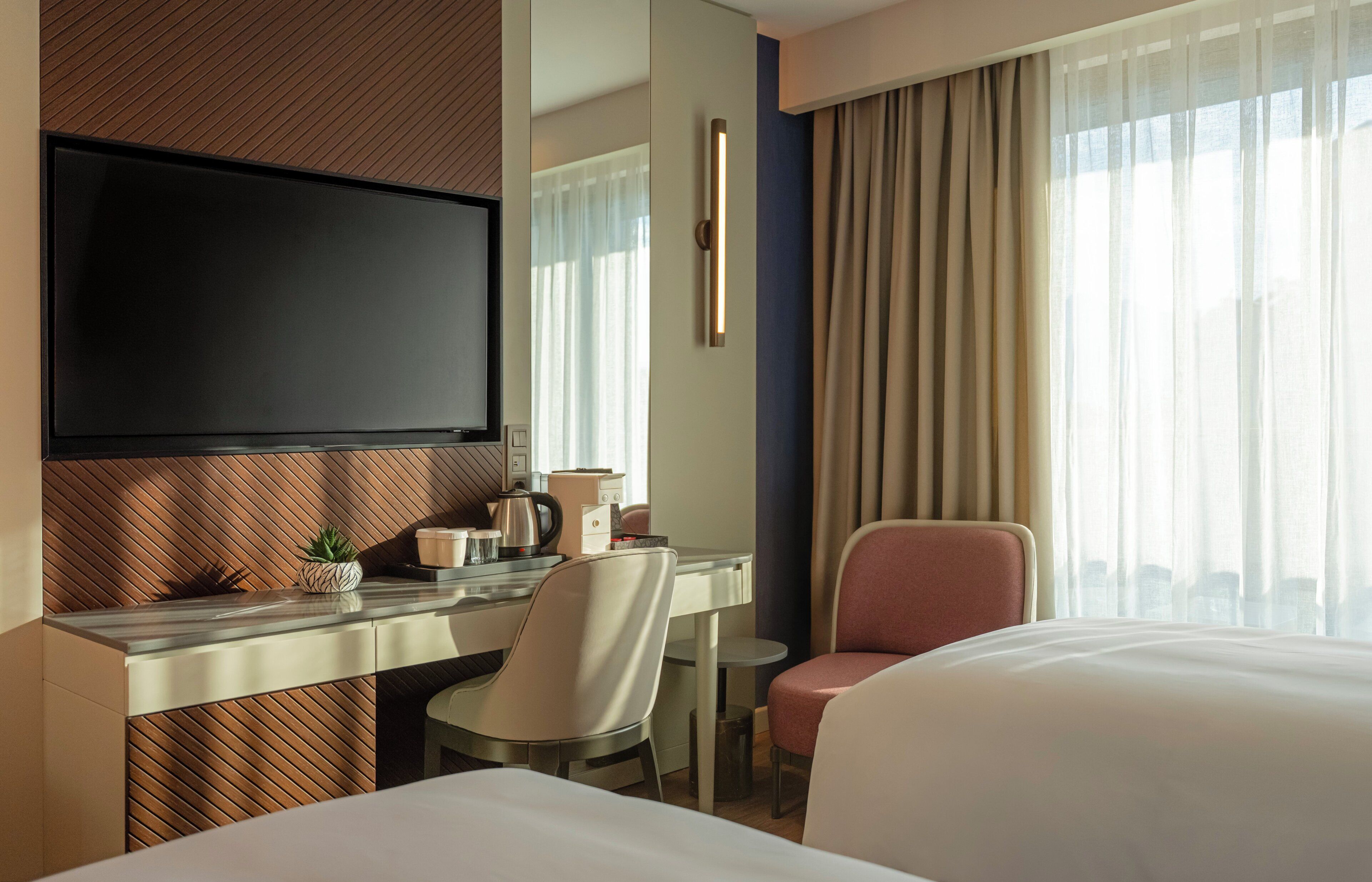 A hotel room with a bed, a desk with a flat-screen TV, a chair, and a window with curtains.
