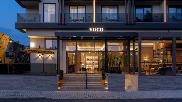 A modern hotel with a glass facade, outdoor seating, and a staircase leading to the entrance.