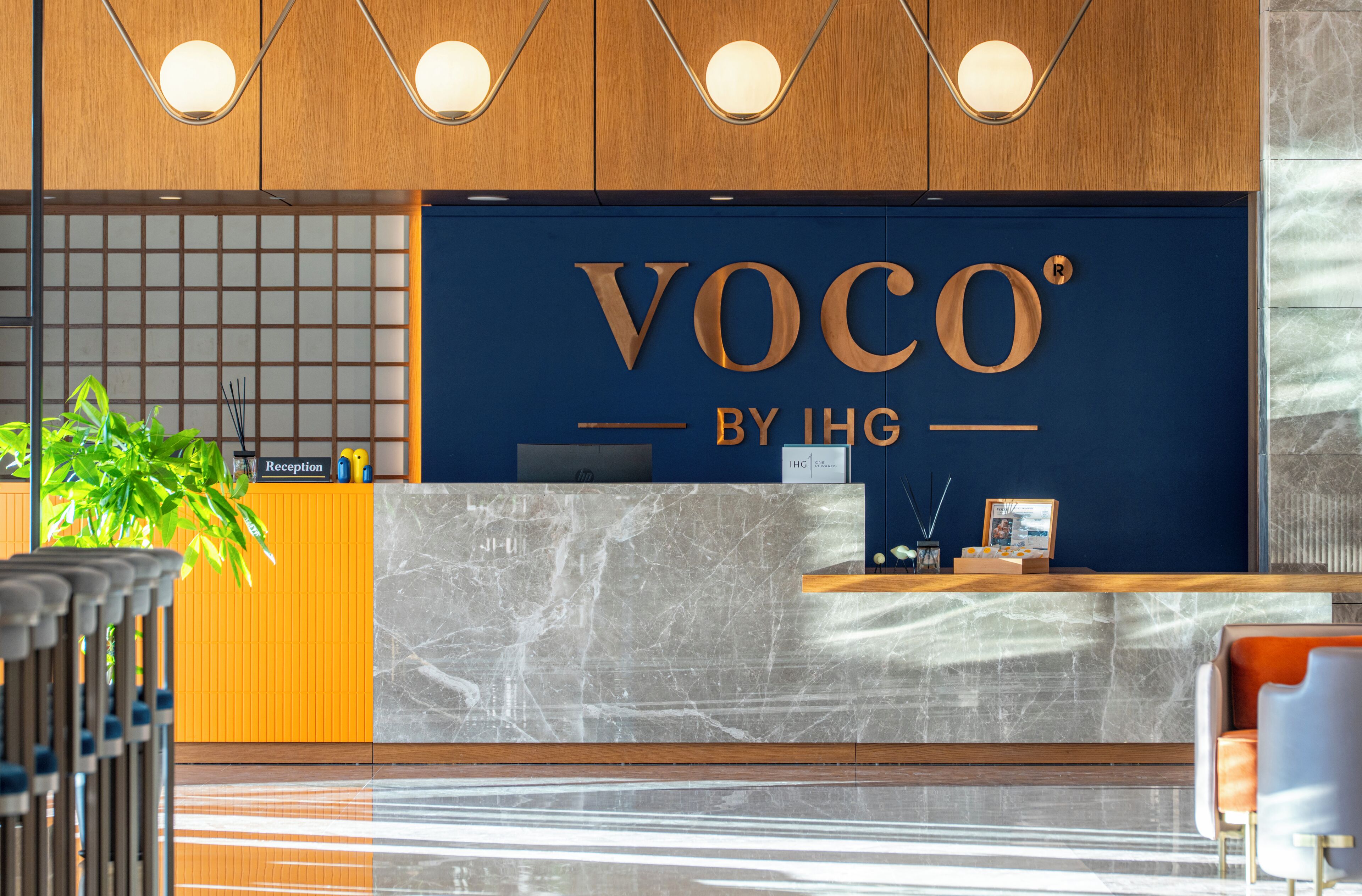 A reception desk with a sign reading 'Voco by IHG' and a reception area with a plant and a framed picture.