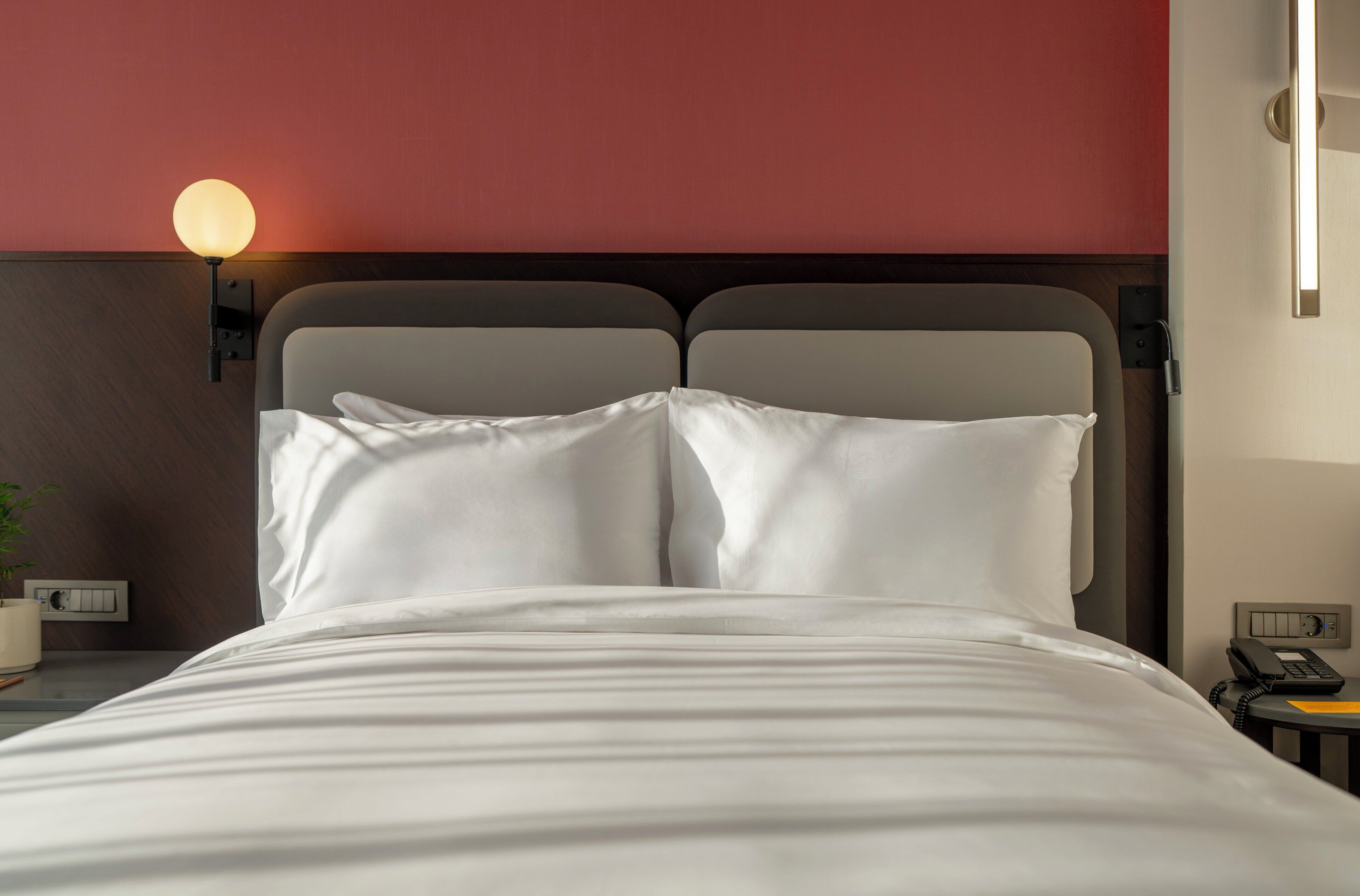 A neatly made bed with white linens, a wall-mounted light fixture, a bedside telephone, and a small table with a plant.
