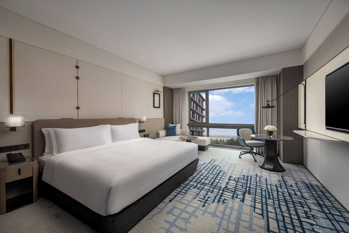 Ein Hotelzimmer mit einem großen Bett, einem Schreibtisch, einem Sessel, einem kleinen Tisch mit einer Vase und einem großen Fenster mit Blick.
