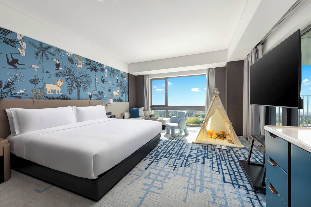 Ein modernes Hotelzimmer mit einem großen Bett, Blick ins Freie, einer kleinen Sitzecke und einem Zelt mit Spielzeug.