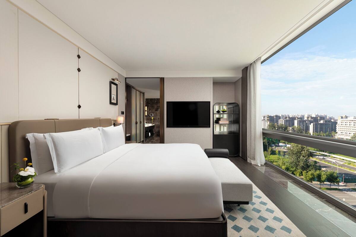 Ein modernes Hotelzimmer mit einem großen Bett, einem Fernseher, einem kleinen Tisch mit Blumen und Blick auf die Stadt durch ein großes Fenster.