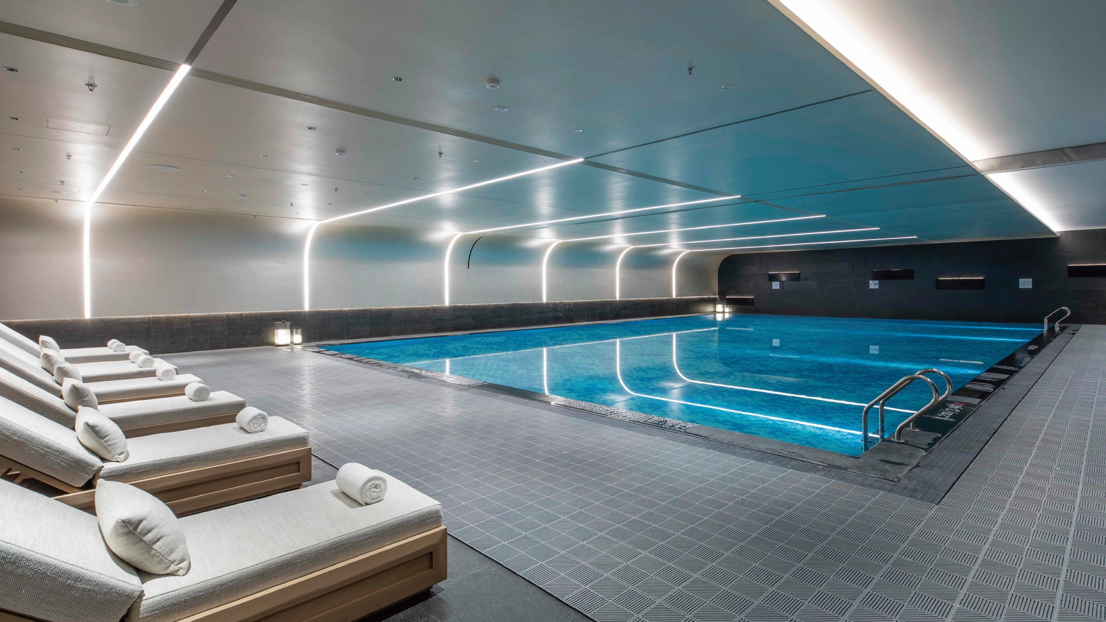 Une piscine intérieure moderne, avec une eau claire et une surface bien dégagée, entourée de chaises longues et de serviettes, et dotée d’un sol à motifs.