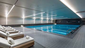 Une piscine intérieure moderne, avec une eau claire et une surface bien dégagée, entourée de chaises longues et de serviettes, et dotée d’un sol à motifs.