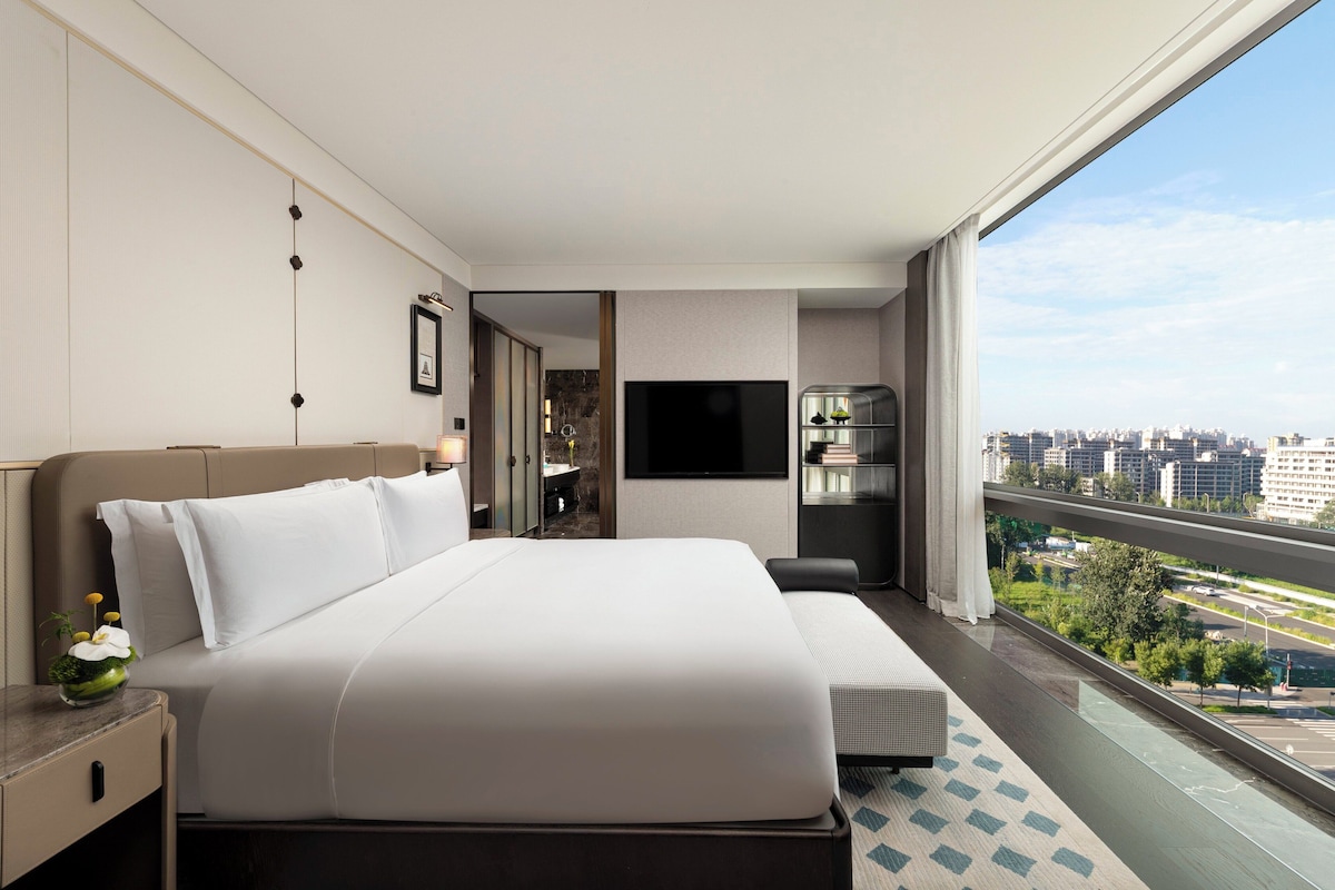Ein modernes Hotelzimmer mit einem großen Bett, einem Fernseher, einem kleinen Tisch mit Blumen und Blick auf die Stadt durch ein großes Fenster.
