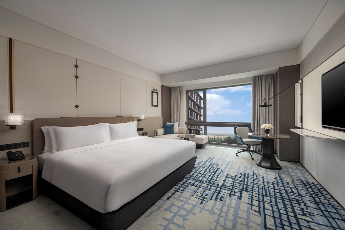 Ein Hotelzimmer mit einem großen Bett, einem Schreibtisch, einem Sessel, einem kleinen Tisch mit einer Vase und einem großen Fenster mit Blick.