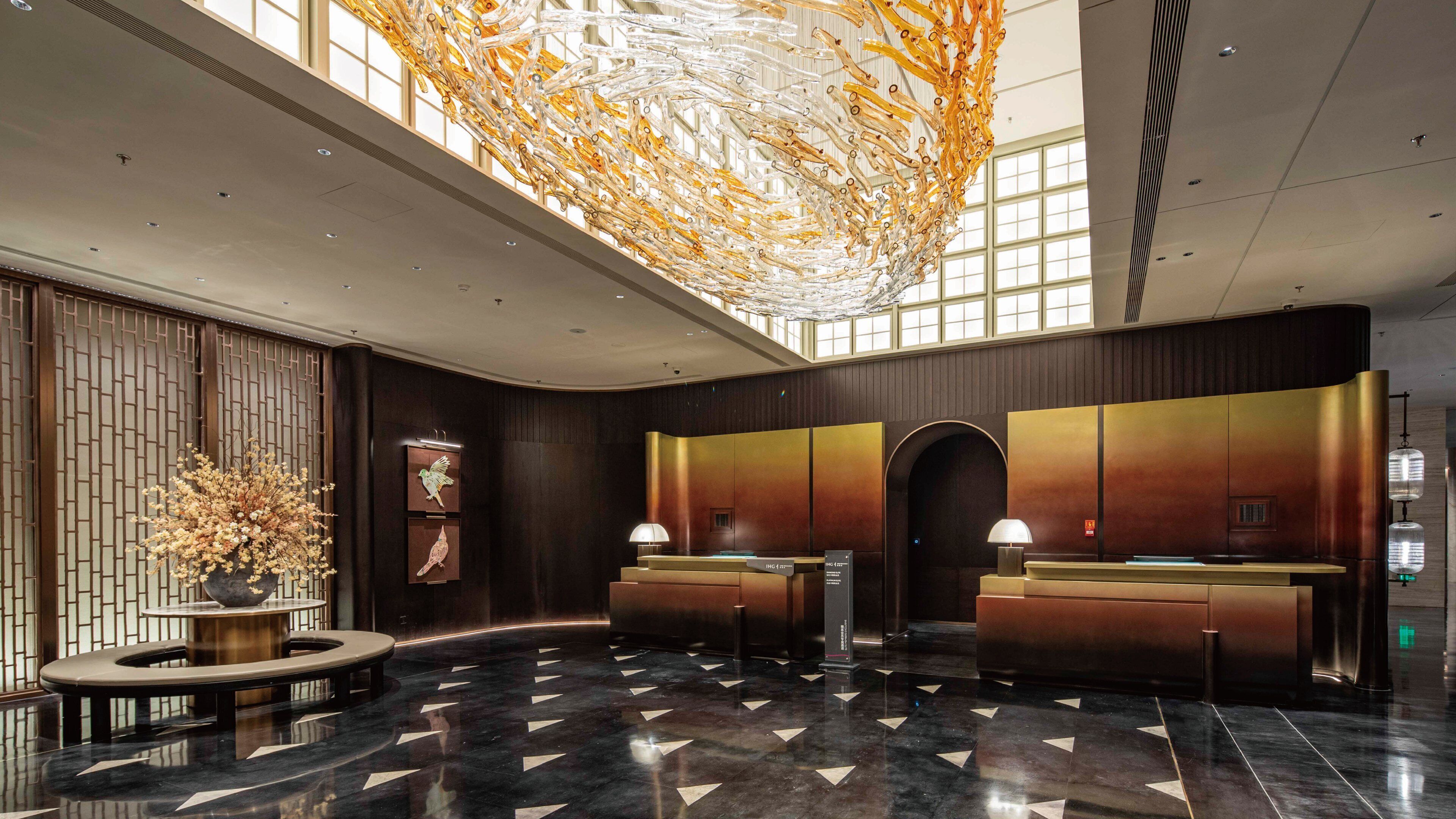 A modern hotel lobby with a reception desk, a decorative ceiling installation, and a large window providing natural light.