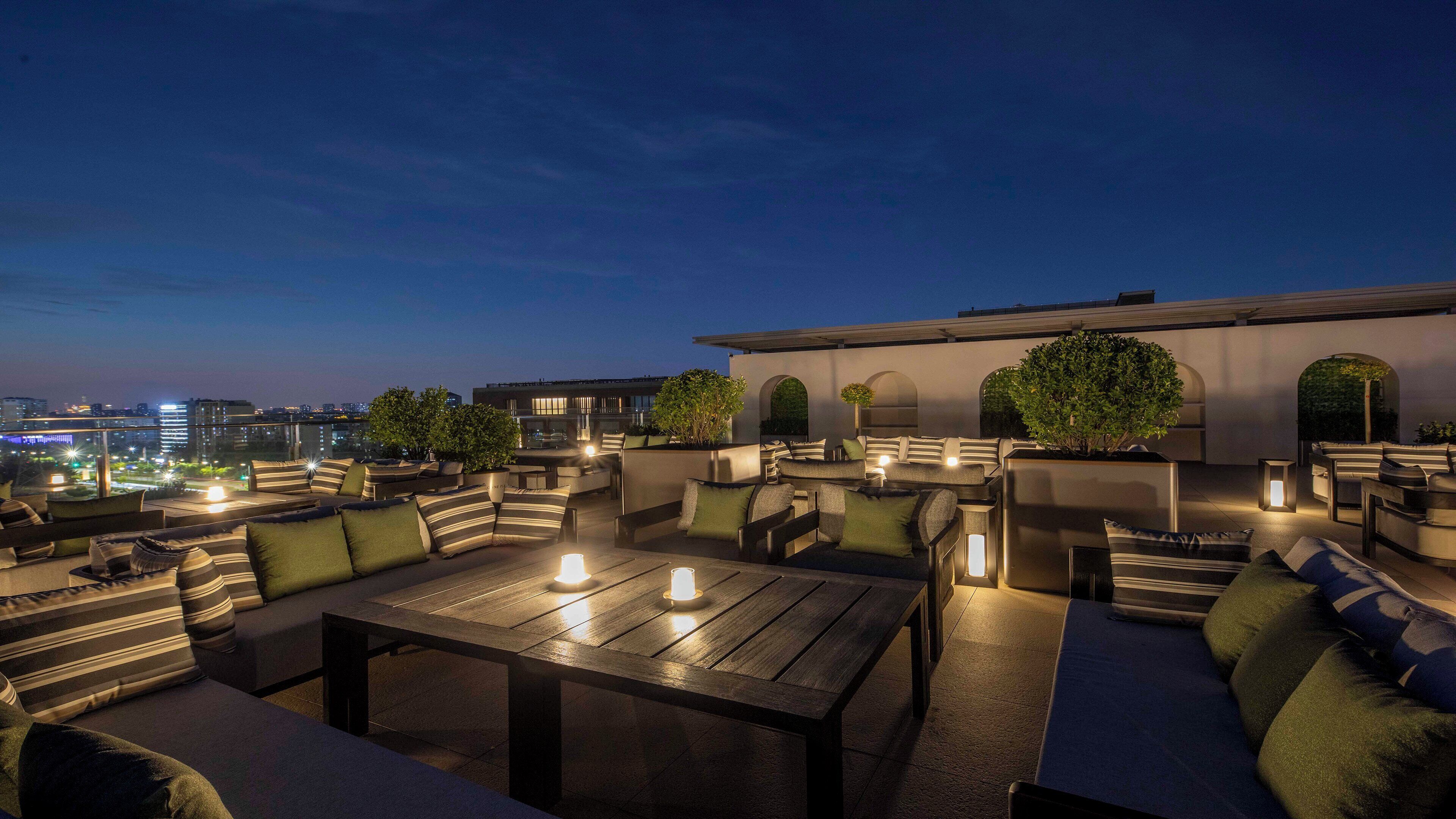 An outdoor seating area with tables and chairs, surrounded by greenery and a cityscape view at dusk.
