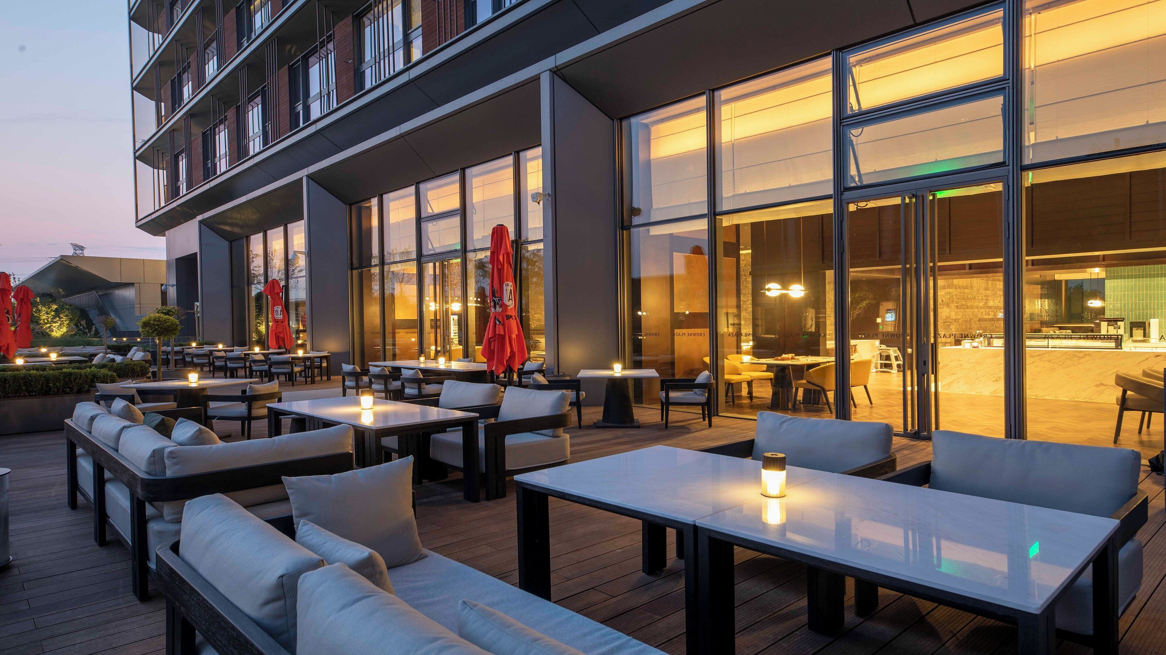 An outdoor seating area with tables and chairs, a building with large windows, and a view of the interior.