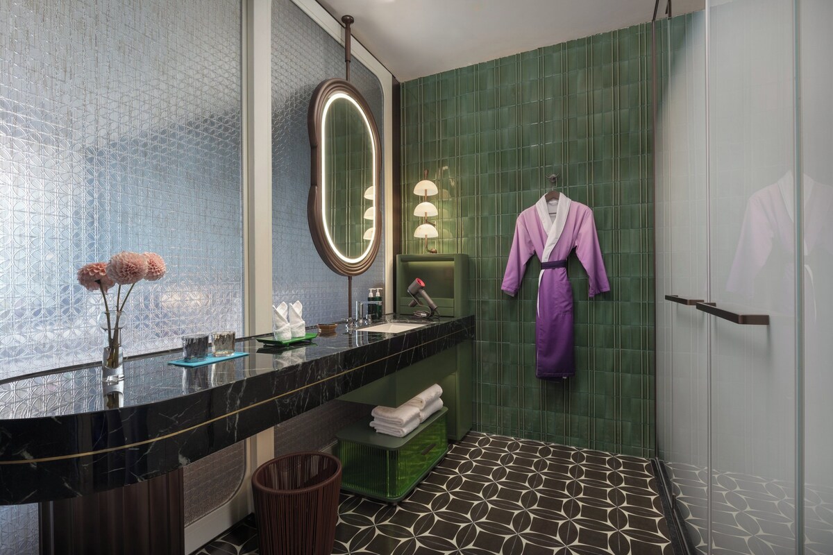 Un baño moderno con encimera de mármol negro, pared de azulejos verdes, cabina de ducha de vidrio y una bata morada colgada en la pared.