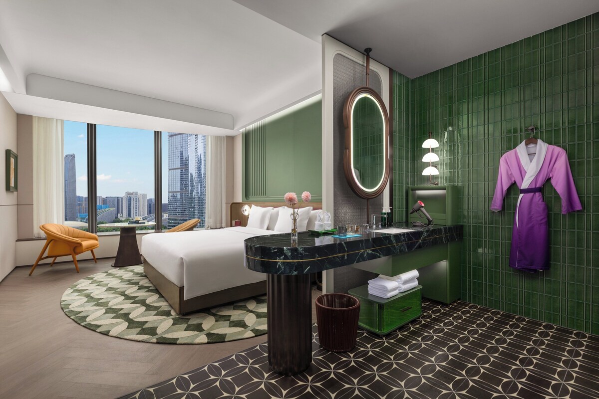 Habitación de hotel con cama, una mesa redonda, baño con pared de azulejos verdes y vista al horizonte de la ciudad.