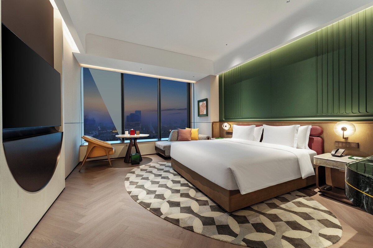 Una habitación de hotel moderna con una cama grande, zona de estar, vistas a la ciudad y un televisor de pantalla plana.