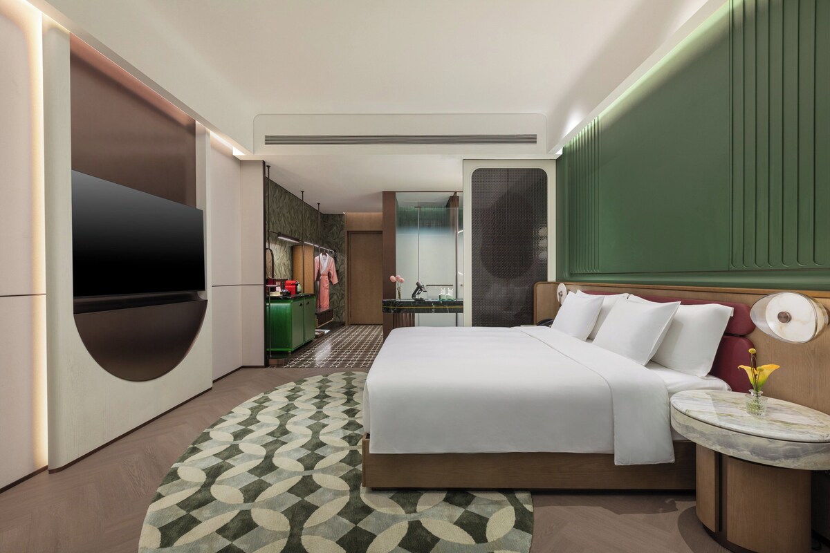 Una habitación de hotel moderna con una cama grande, un televisor, una mesita con un florero y una pared verde decorativa.