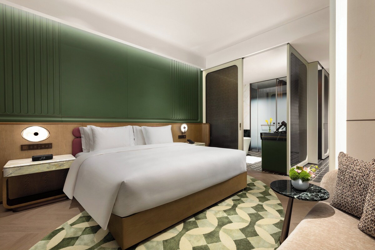 Una habitación de hotel moderna con una cama grande, una mesita de noche, una pequeña mesa con un florero y una pared de acento verde.