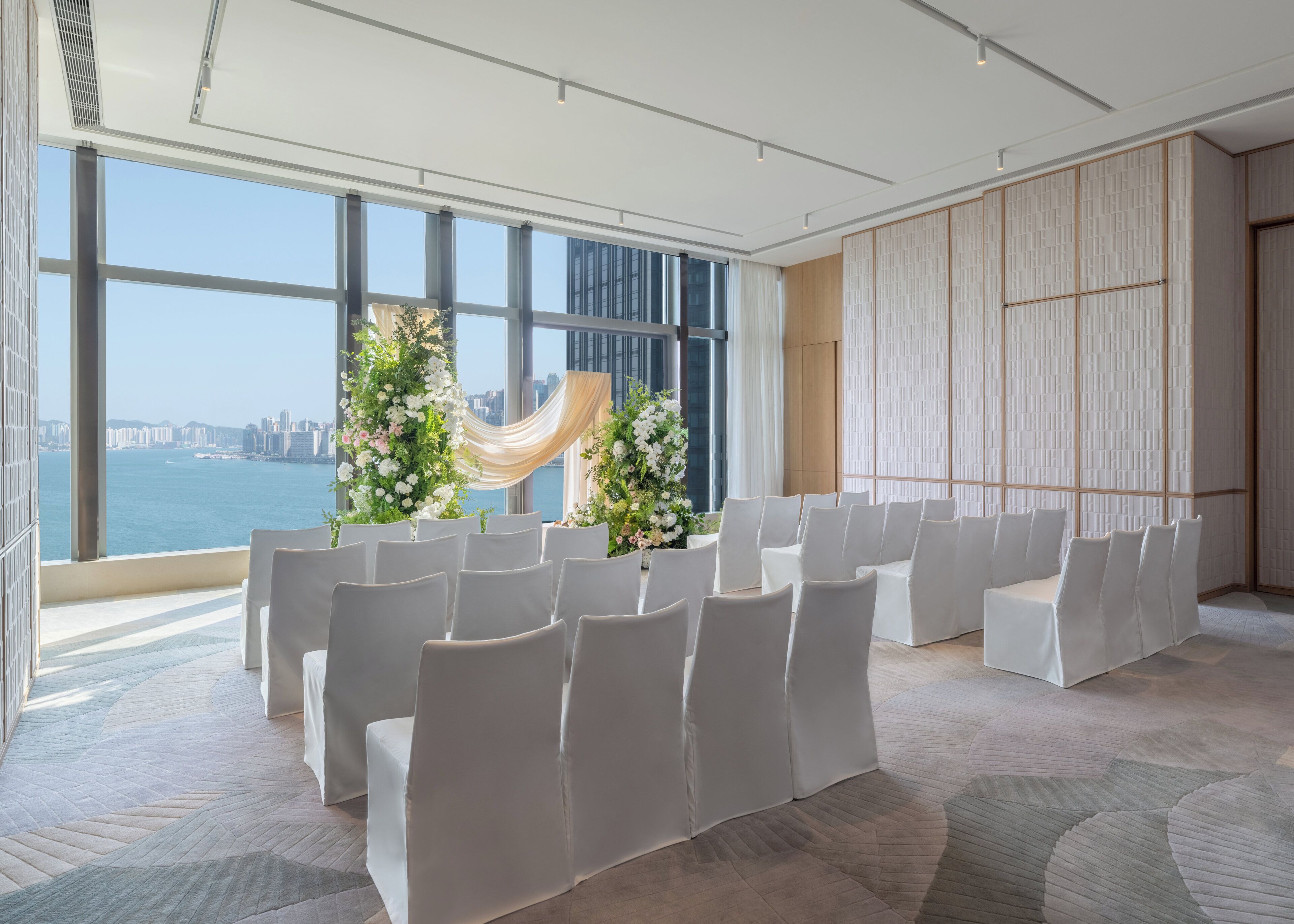 Un salón de conferencias con hileras de sillas blancas, amplias ventanas con vista a la ciudad y un arreglo floral decorativo.
