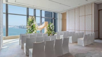 Un salón de conferencias con hileras de sillas blancas, amplias ventanas con vista a la ciudad y un arreglo floral decorativo.
