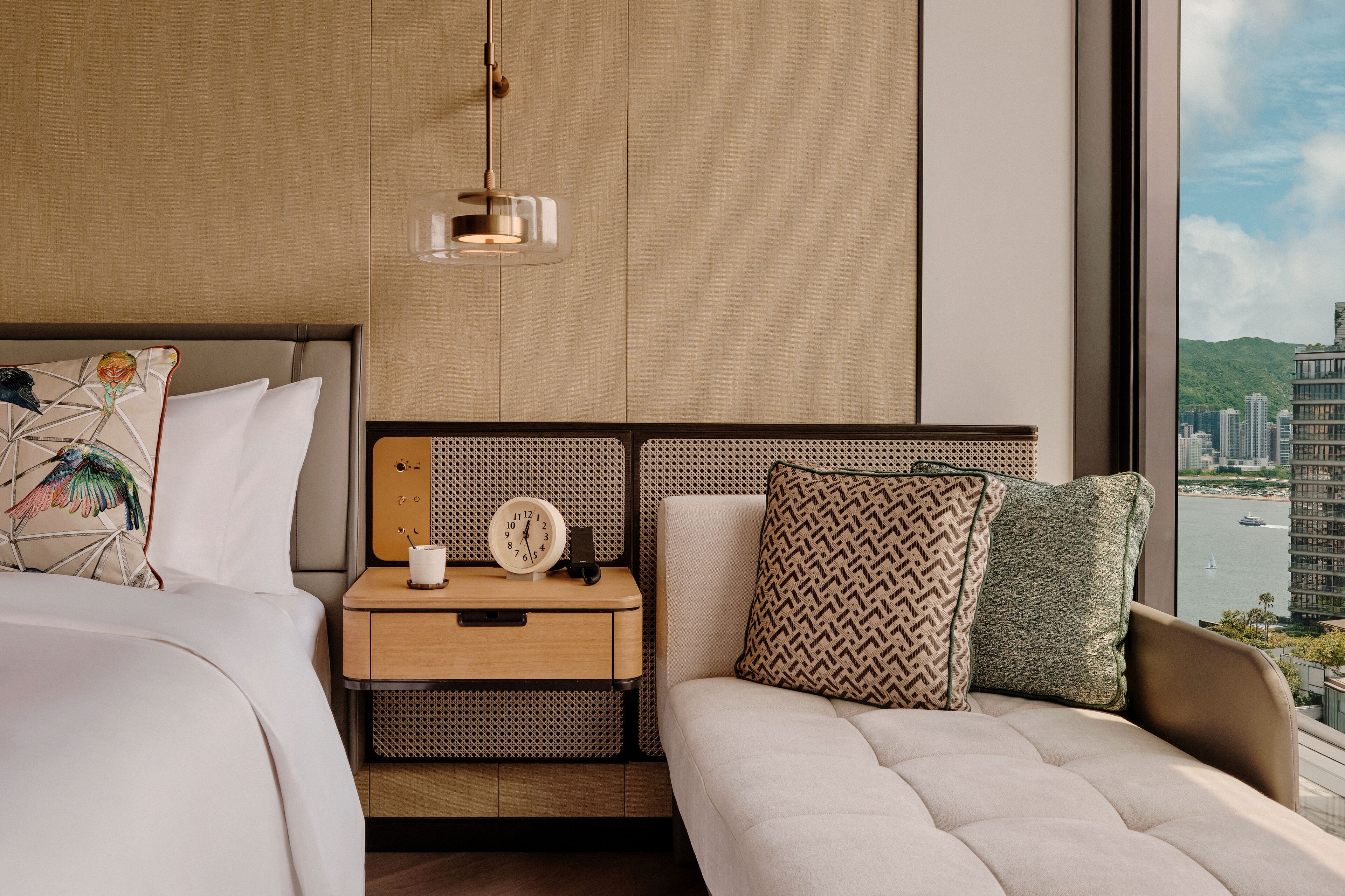 Et moderne hotelværelse med en stor seng, et sengebord med et ur, en lampe og udsigt over en by gennem et stort vindue.