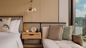 A modern hotel room with a large bed, a bedside table with a clock, a lamp, and a view of a cityscape through a large window.
