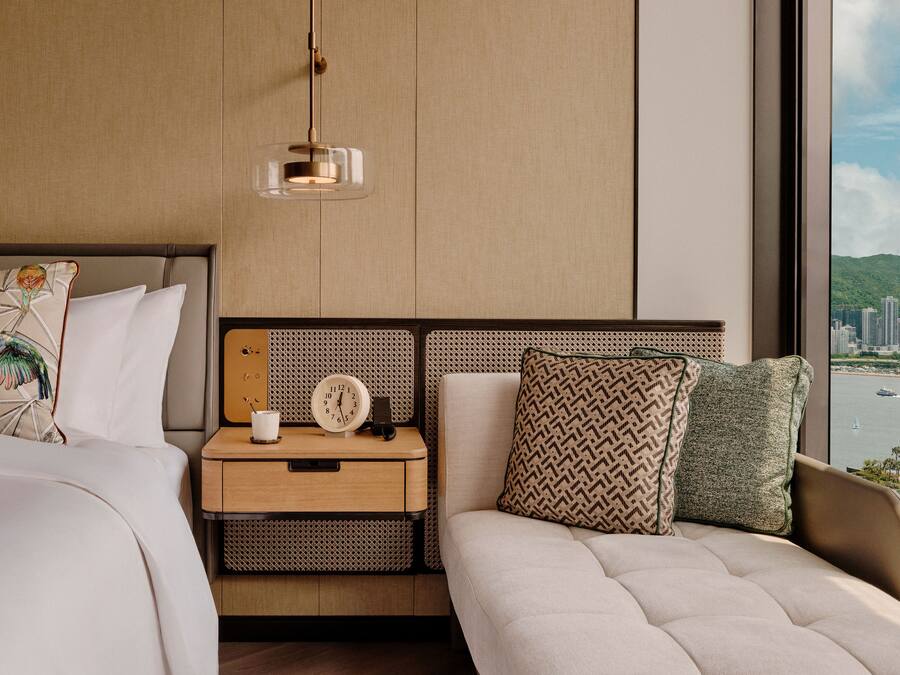 A modern hotel room with a large bed, a bedside table with a clock, a lamp, and a view of a cityscape through a large window.