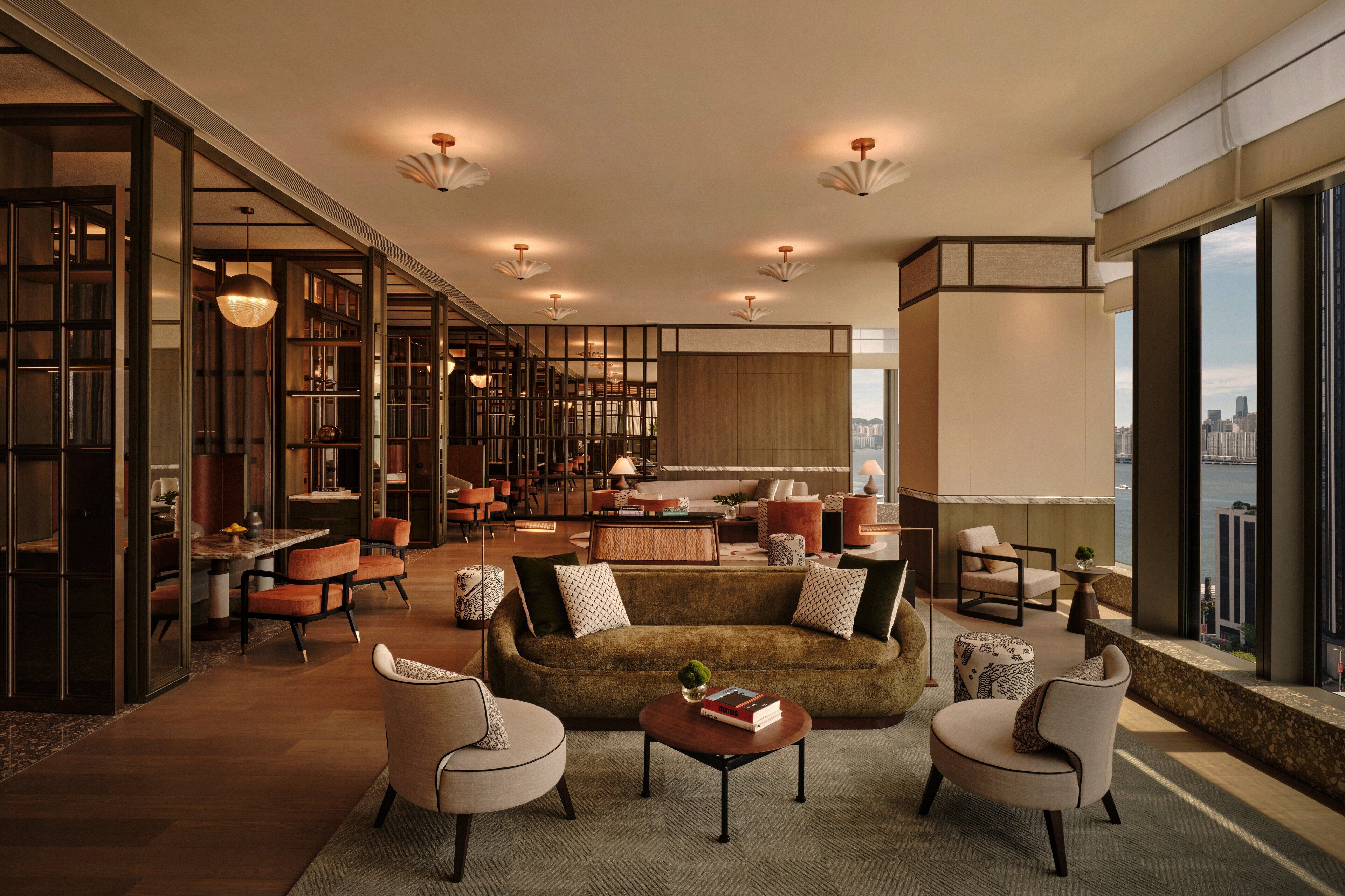 Un vaste espace de vie comprenant un canapé, des fauteuils et une table basse. On aperçoit une cuisine au fond et de grandes fenêtres qui offrent une vue sur la ville.
