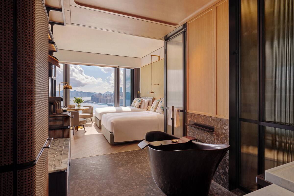 Ein modernes Hotelzimmer mit einem großen Bett, einer Sitzecke mit Tisch und Stühlen, einer Badewanne und Blick auf die Stadt durch große Fenster.