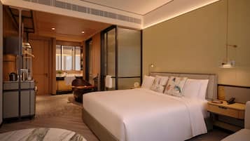 A modern hotel room with a large bed, a desk, a chair, a wardrobe, and a bathroom with a glass door.