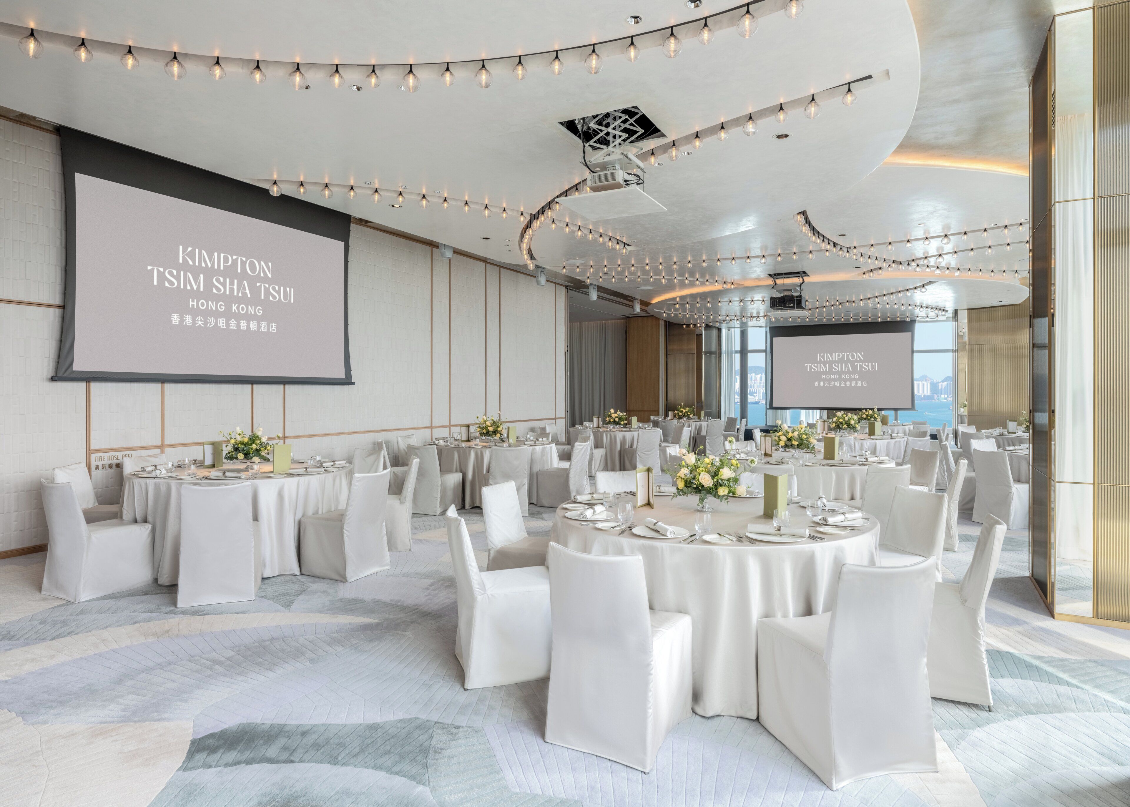 A conference room with multiple tables, chairs, and a large screen displaying text.