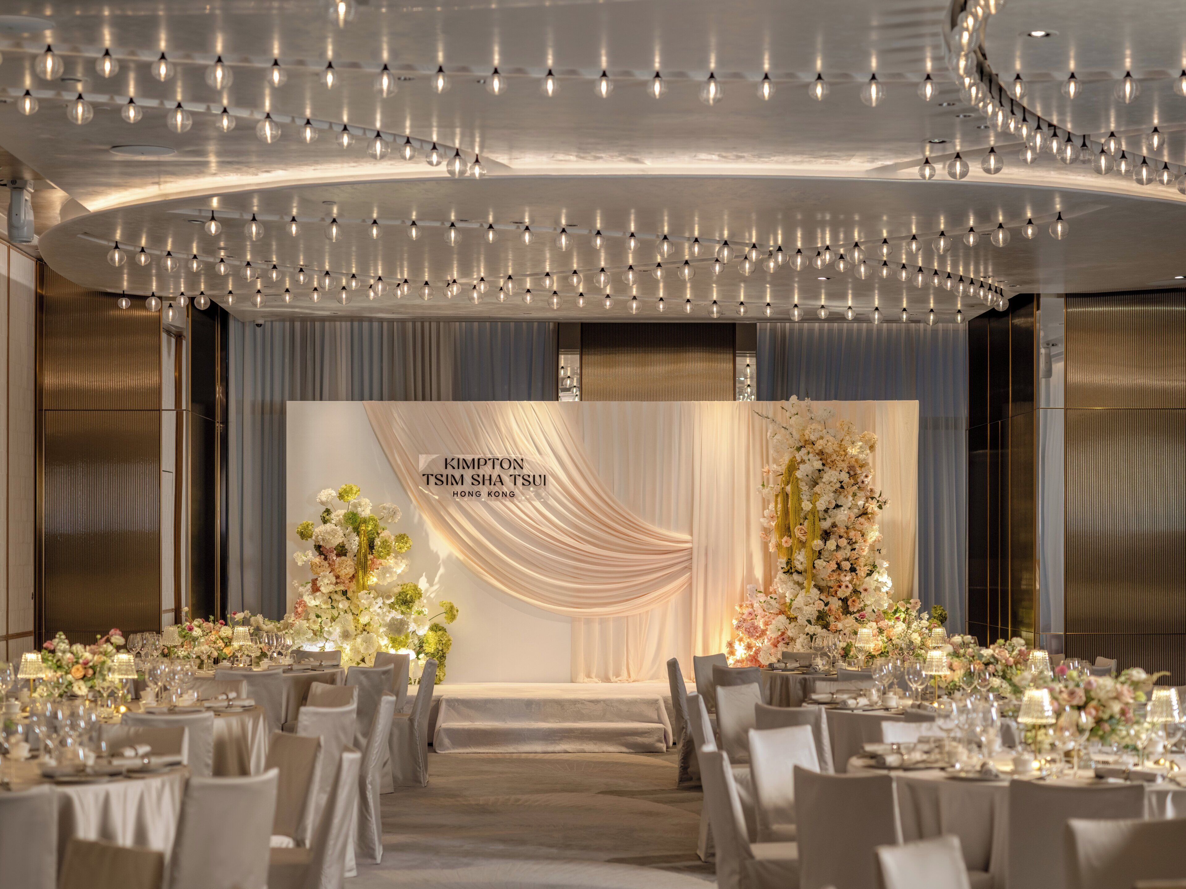 Une salle de banquet dotée d’une scène, de tables dressées pour le repas et d’un décor sur lequel figure le nom « KIMPTON TSIM SHA TSUI HONG KONG ».