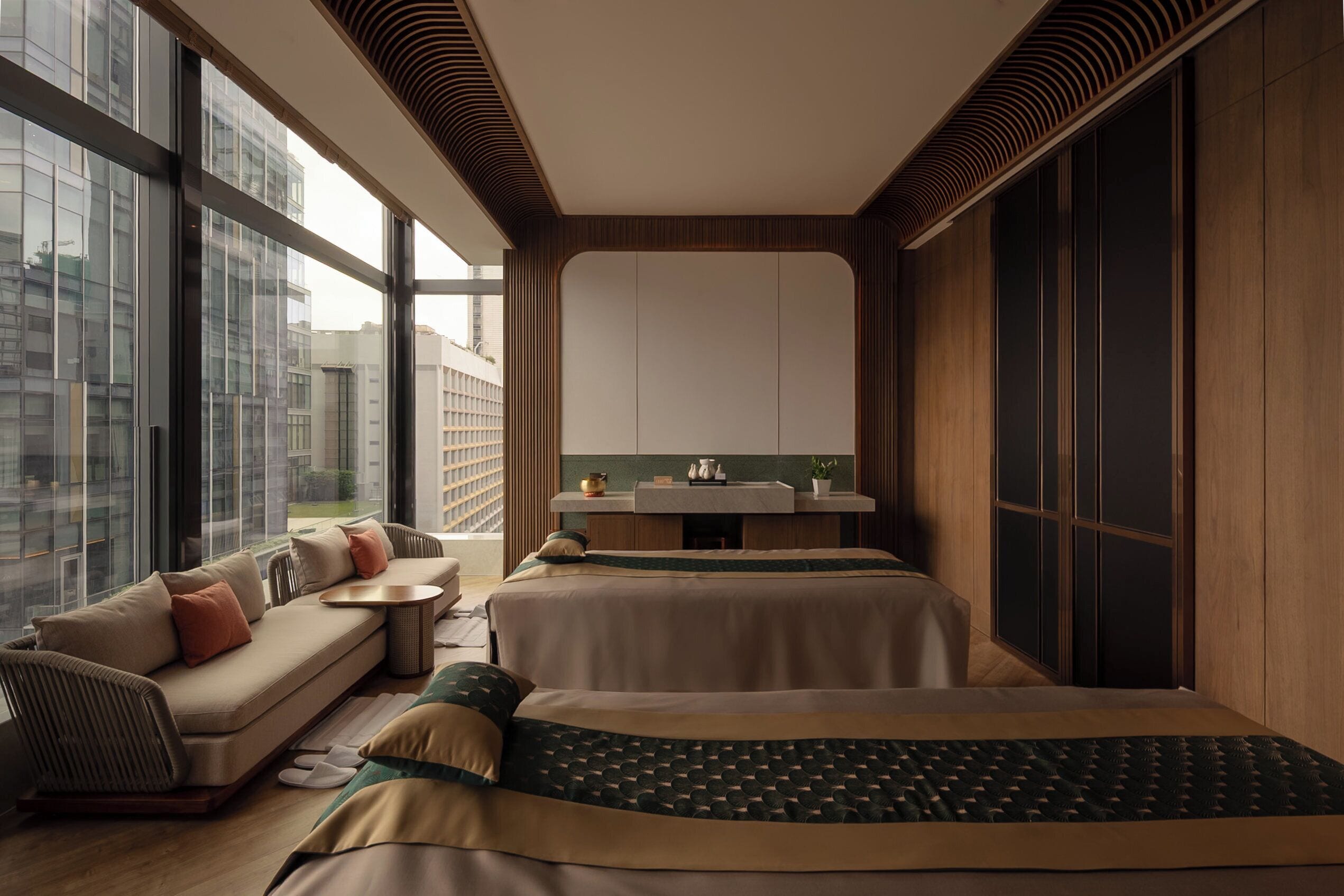 A modern hotel room with a sofa, two beds, a small table, and a view of the city.
