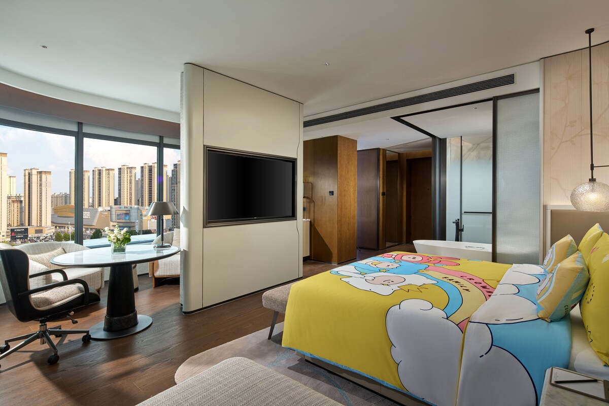 Una habitació d'hotel moderna amb una finestra gran, un llit amb un llençol estampat amb motius de núvols, una taula de treball amb una cadira i un televisor de pantalla plana.