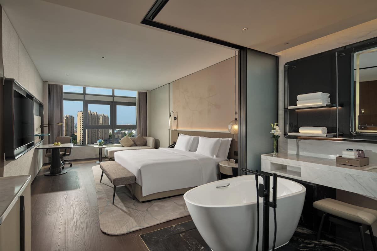 A modern hotel room with a large bed, a sitting area, a large bathtub, a vanity with a mirror, and a television.