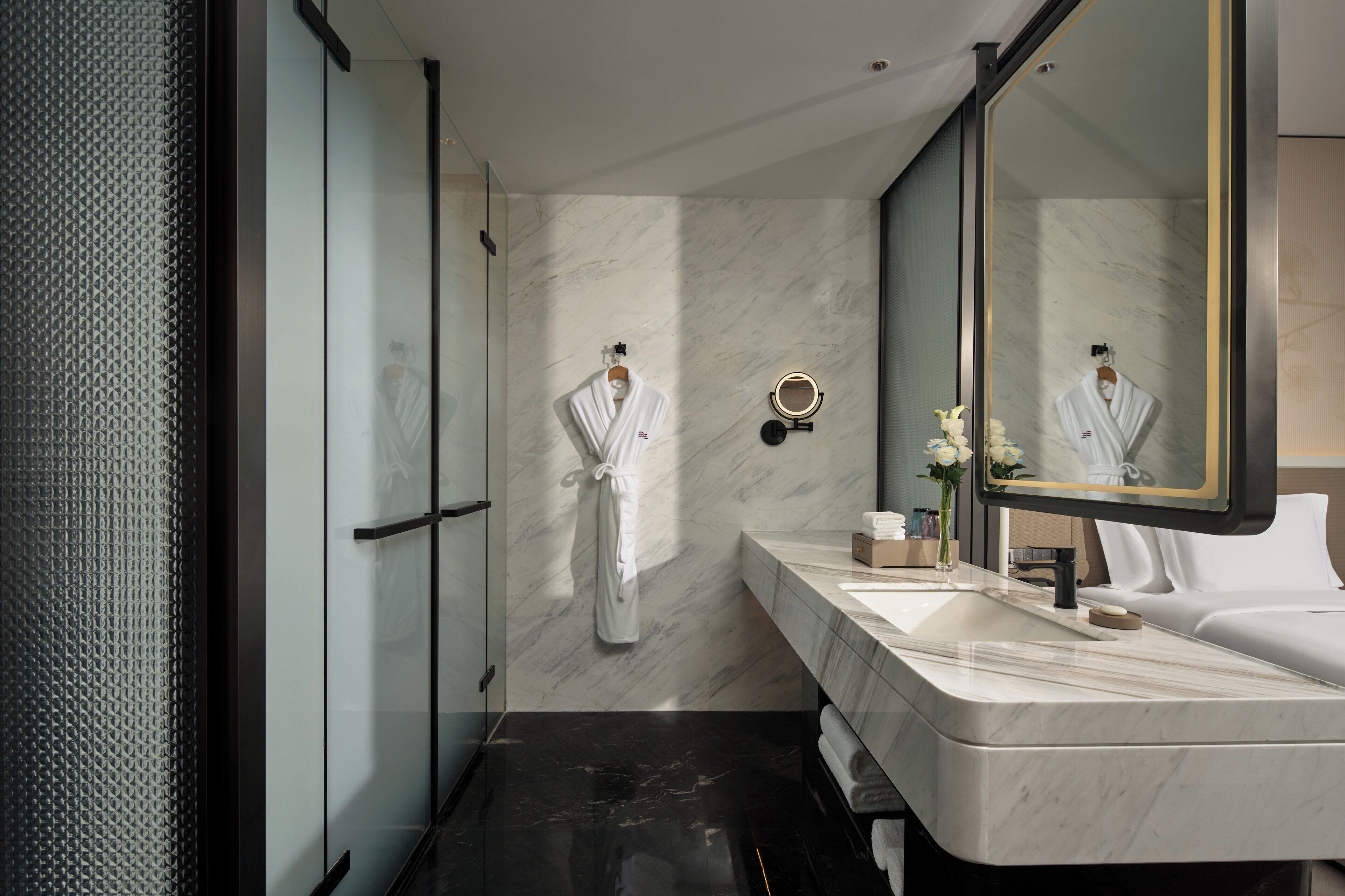 A modern bathroom with a large mirror, a white robe hanging, a marble sink, and a black floor.