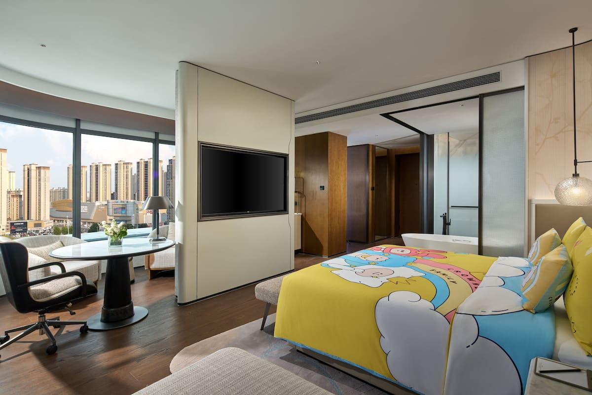 A modern hotel room with a large window, a bed with a colorful cloud-themed bedspread, a desk with a chair, and a flat-screen TV.